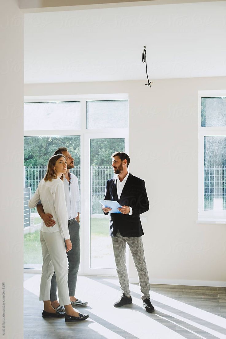 Real estate agent showing a modern, sunlit room to a couple standing near large windows.