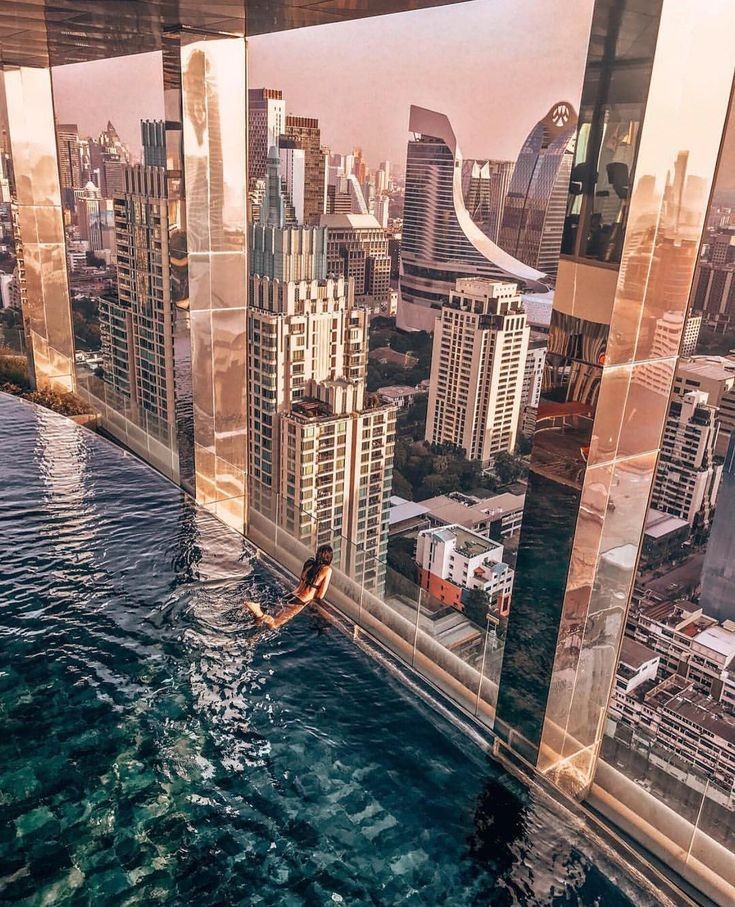 Person swimming in an infinity pool overlooking a city skyline at sunset with modern high-rise buildings.