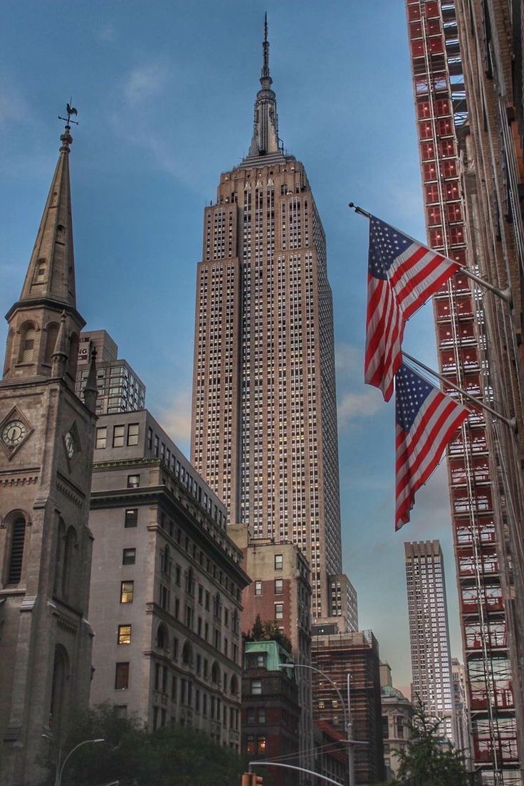 View of the Empire State Building with surrounding buildings and American flags, under a clear blue sky.